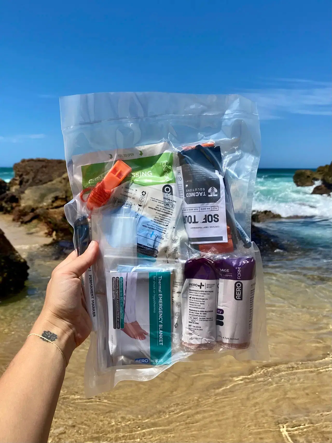 A sealed kit insert held up on the beach, showing the tourniquet, dressings and blanket inside