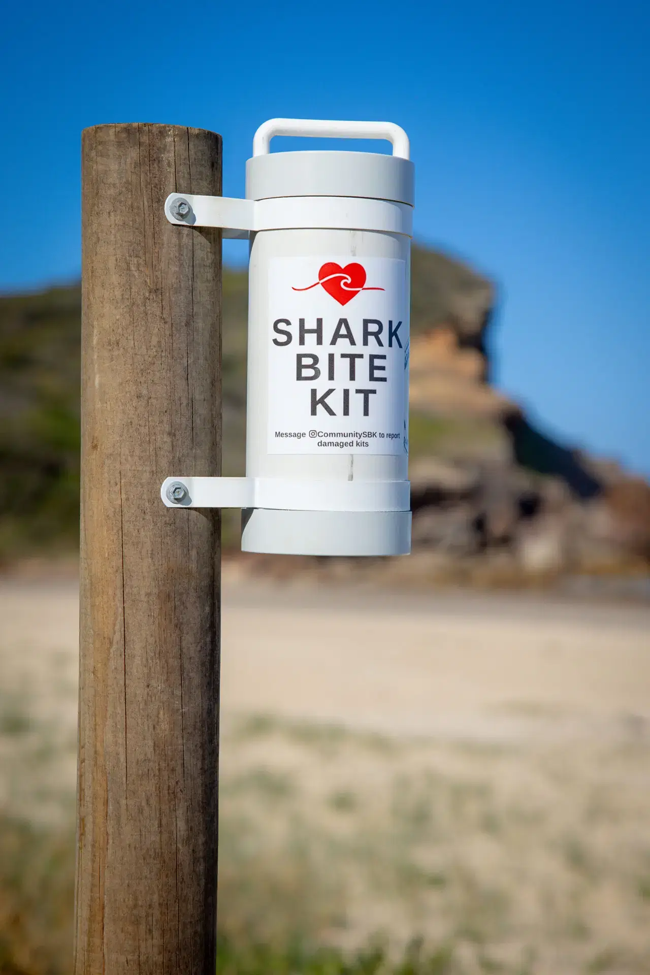 A Shark Bite Kit mounted on a timber post at a beach access track, headland behind